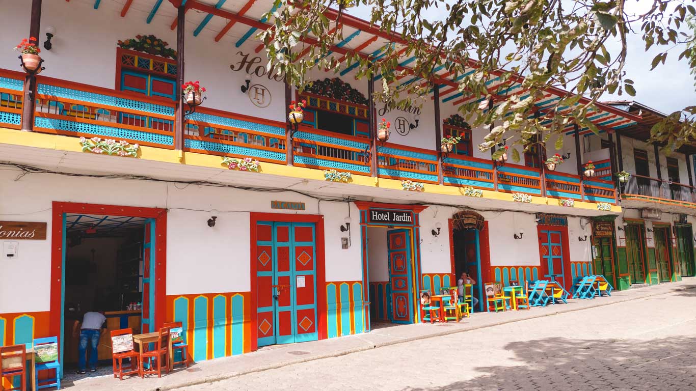 Una fachada colorida de estilo tradicional en el pueblo de Jardín, Colombia, con balcones decorados y macetas con flores. El edificio alberga el "Hotel Jardín", con puertas y ventanas pintadas en tonos vivos de rojo, azul y amarillo. En la parte inferior, hay sillas y mesas en la acera, añadiendo un ambiente acogedor y pintoresco al lugar. Un hombre se inclina dentro de uno de los establecimientos, y algunas personas se sientan en las mesas disfrutando del entorno.