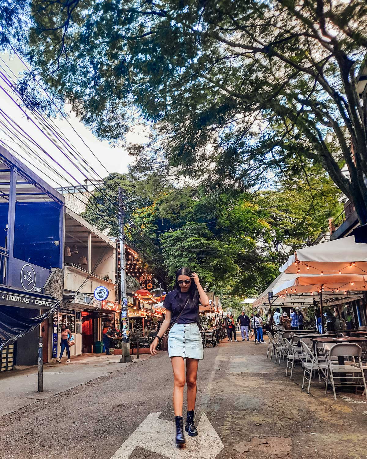 Una mujer camina por una calle bordeada de árboles en una zona peatonal, vestida con una falda de mezclilla y una camiseta negra, usando gafas de sol. A su alrededor hay restaurantes y bares con luces colgantes y mesas al aire libre, creando un ambiente animado y acogedor. El entorno muestra un lugar vibrante y lleno de vida, con algunas personas caminando al fondo.