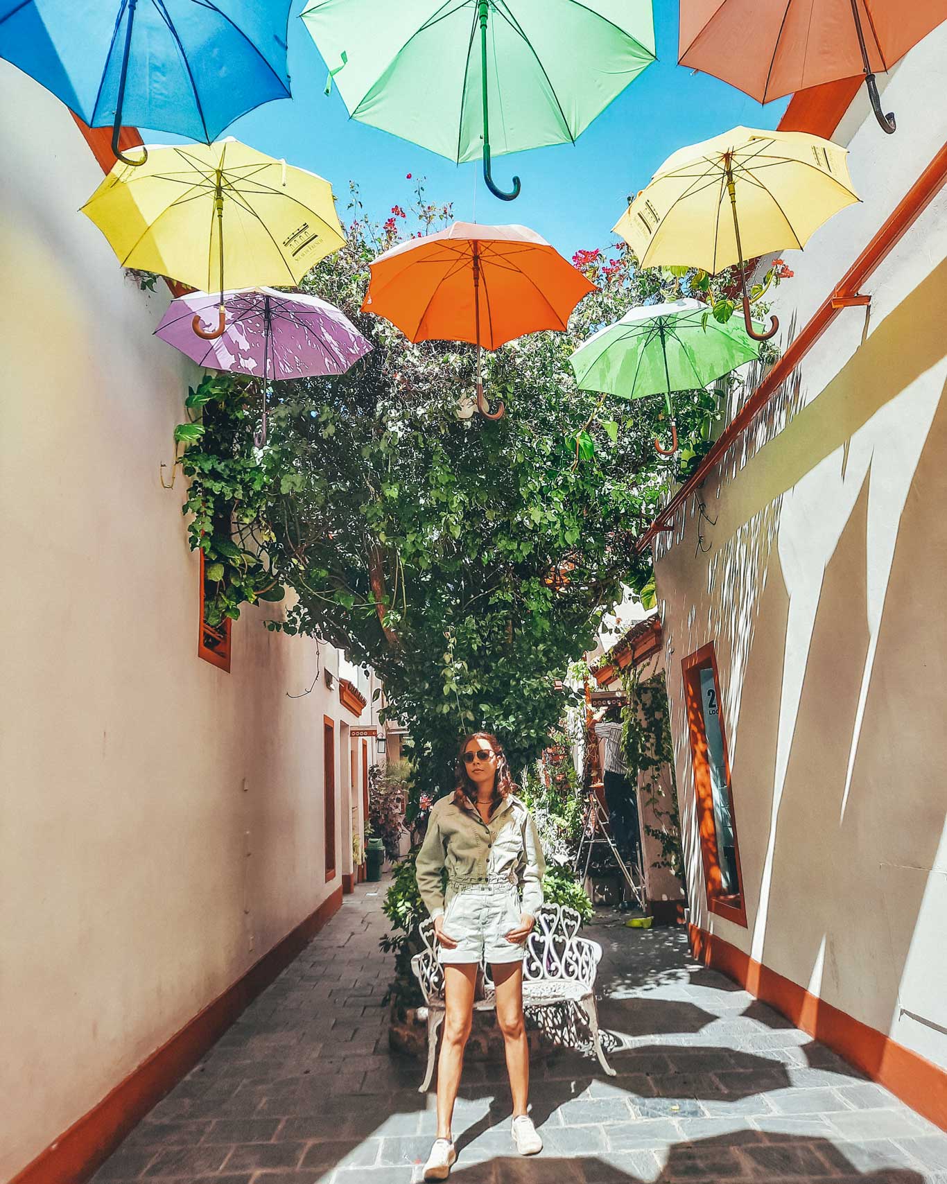 Una persona de pie en un pasaje estrecho al aire libre, decorado con coloridos paraguas suspendidos en el aire. Las paredes del pasaje son claras, y se observan plantas y vegetación alrededor, añadiendo un ambiente fresco y vibrante. La persona está en el centro, mirando hacia la cámara, bajo la sombra proyectada por los paraguas y la vegetación.