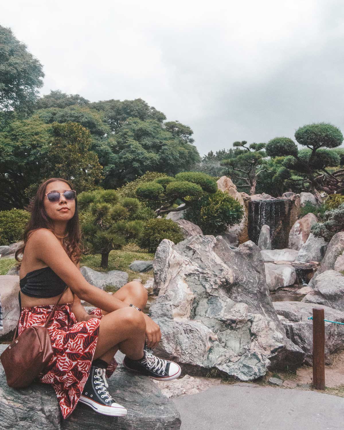 Una persona sentada sobre una roca en el Jardín Japonés de Buenos Aires, Argentina, rodeada de árboles podados y vegetación típica de estilo japonés. Al fondo se aprecia una pequeña cascada que fluye entre las piedras, creando un ambiente sereno y natural. El cielo está nublado, realzando el tono tranquilo del lugar.