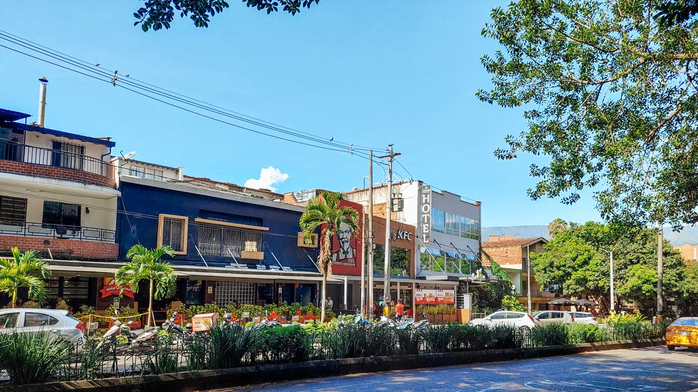 Una calle en el barrio Laureles, Medellín, con una serie de edificios comerciales y residenciales. Se destaca un restaurante KFC con su icónico logotipo del coronel Sanders en la fachada. El entorno está rodeado de árboles y plantas y se ven algunos coches estacionados frente a los negocios. El cielo está despejado, dando un ambiente soleado y agradable a la escena. El barrio es una de las mejores opciones dónde alojarse en Medellín.