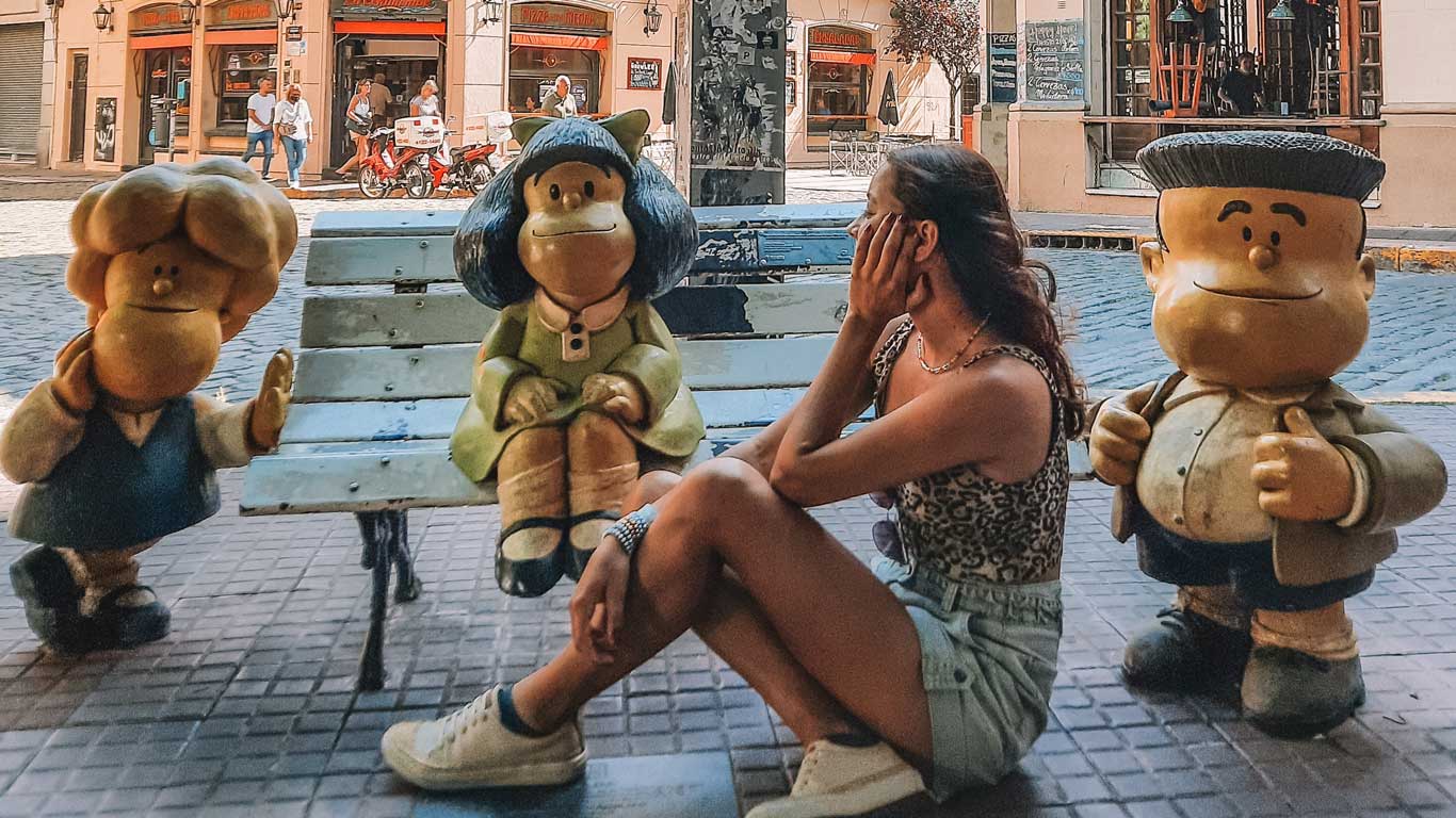 Una persona sentada junto a estatuas de los personajes de Mafalda en una calle adoquinada. Las figuras incluyen a Mafalda y sus amigos, que están colocados alrededor de un banco, creando una escena amigable y nostálgica. Al fondo, se observa una calle con tiendas y transeúntes, añadiendo un ambiente urbano típico de San Telmo, Buenos Aires.