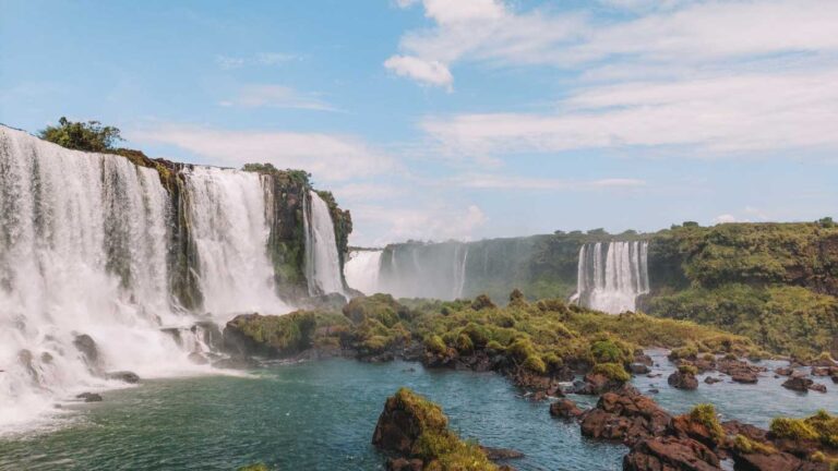 La imagen muestra las majestuosas Cataratas del Iguazú, un conjunto impresionante de cascadas rodeadas de vegetación exuberante. El agua cae con fuerza sobre formaciones rocosas, creando una niebla que se eleva al cielo azul con algunas nubes dispersas. A la derecha, se pueden ver otras cascadas más distantes en medio del paisaje verde, característico de la región subtropical.