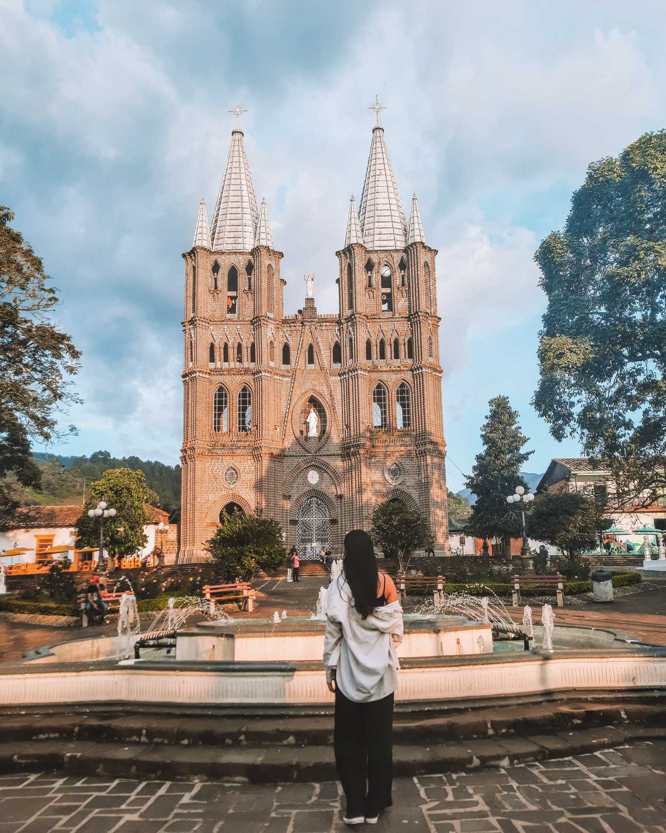 Una mujer de espaldas, vestida con una camisa holgada blanca y pantalones oscuros, observa una imponente catedral de estilo neogótico en el pueblo de Jardín, Colombia. La iglesia tiene dos altas torres simétricas y está construida con ladrillos marrones, con una estatua blanca de la Virgen María en el centro. Frente a la iglesia, hay una fuente rodeada de bancos y vegetación. El cielo es azul con algunas nubes, añadiendo un toque tranquilo a la escena.