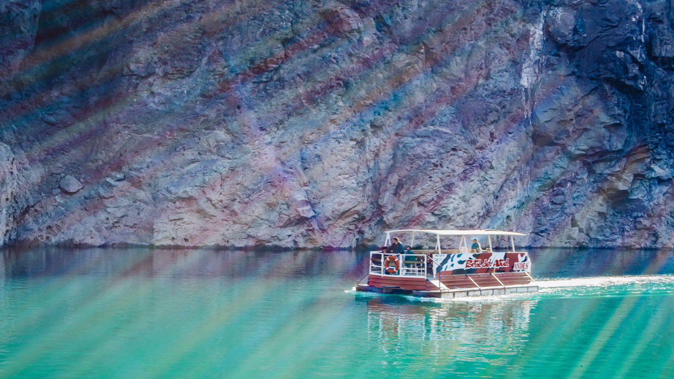 La imagen muestra una lancha turística navegando por aguas turquesas en un lago rodeado de paredes rocosas. La luz del sol crea un efecto de rayos diagonales sobre la escena. El entorno es montañoso y la embarcación lleva a un grupo de personas disfrutando del paisaje natural, típico de la región de San Rafael, Mendoza.