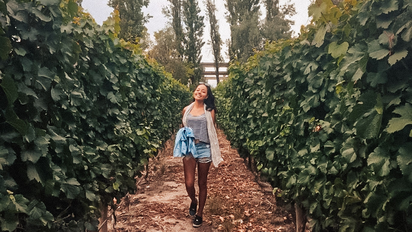 La imagen muestra a una mujer caminando alegremente entre los viñedos, rodeada de altas hileras de vides verdes. Viste un conjunto casual de verano con una camiseta a rayas y shorts, mientras sostiene una chaqueta ligera en la mano. El paisaje refleja un entorno natural y sereno, típico de las regiones vinícolas de Mendoza.