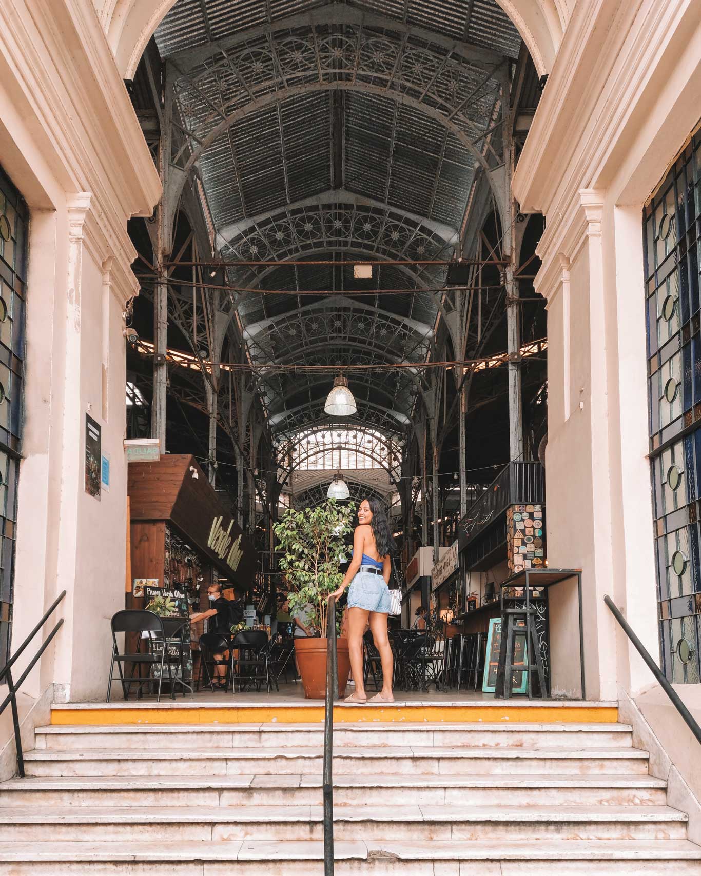 Una persona posando en la entrada del mercado de San Telmo cubierto con una estructura de hierro y un techo alto de estilo industrial. El mercado tiene varios locales visibles al fondo, con mesas y sillas dispuestas en su interior. La persona está de pie en la parte superior de unas escaleras, sonriendo hacia la cámara, con plantas y detalles arquitectónicos a su alrededor.