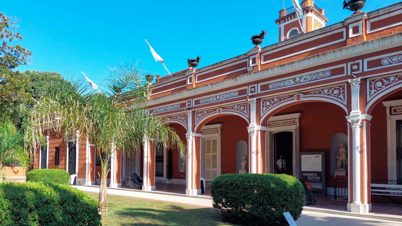 Imagen de la fachada de un edificio de estilo colonial con arcos decorados, columnas blancas y detalles ornamentales en tonos rojizos. El edificio está rodeado de vegetación, con palmeras y arbustos bien cuidados en primer plano, bajo un cielo azul claro.