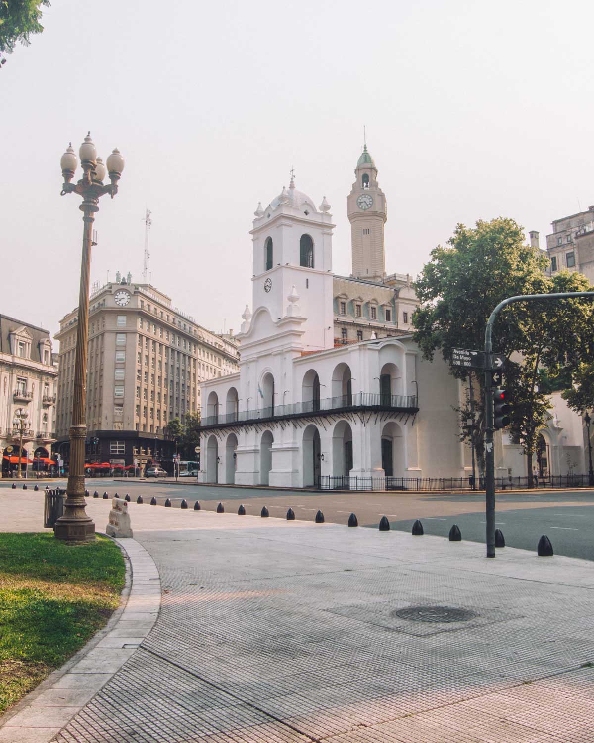 Imagen del Cabildo de Buenos Aires, Argentina, con su distintiva fachada blanca y torre con reloj. El entorno muestra edificios históricos y un farol de estilo antiguo en primer plano. La plaza se ve tranquila, con una mezcla de áreas pavimentadas y verdes alrededor del edificio.