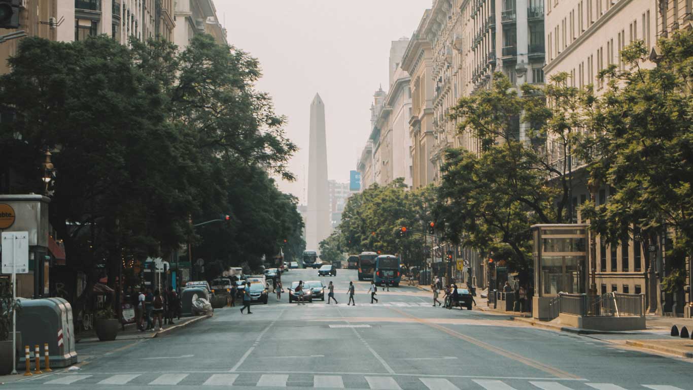 Vista de una calle en Buenos Aires, Argentina, con el Obelisco al fondo en el centro de la imagen. La calle está bordeada por edificios altos y árboles frondosos, mientras personas cruzan por el paso de peatones. El ambiente urbano está animado con tráfico y una mezcla de vehículos y peatones. Caminar en el centro es una de las mejores cosas que hacer en Buenos Aires en 3 días.