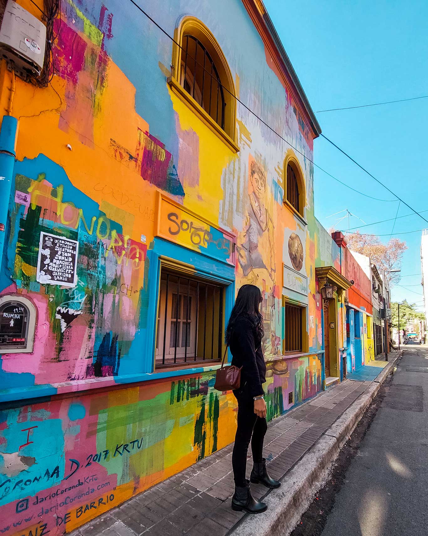 Una persona de pie frente a una fachada vibrante y colorida en el barrio de Palermo, Buenos Aires, Argentina. El edificio está cubierto de murales y grafitis con una mezcla de tonos vivos como el amarillo, azul, rosa y verde, creando una atmósfera artística y moderna. El cielo despejado resalta los colores brillantes de la escena urbana.