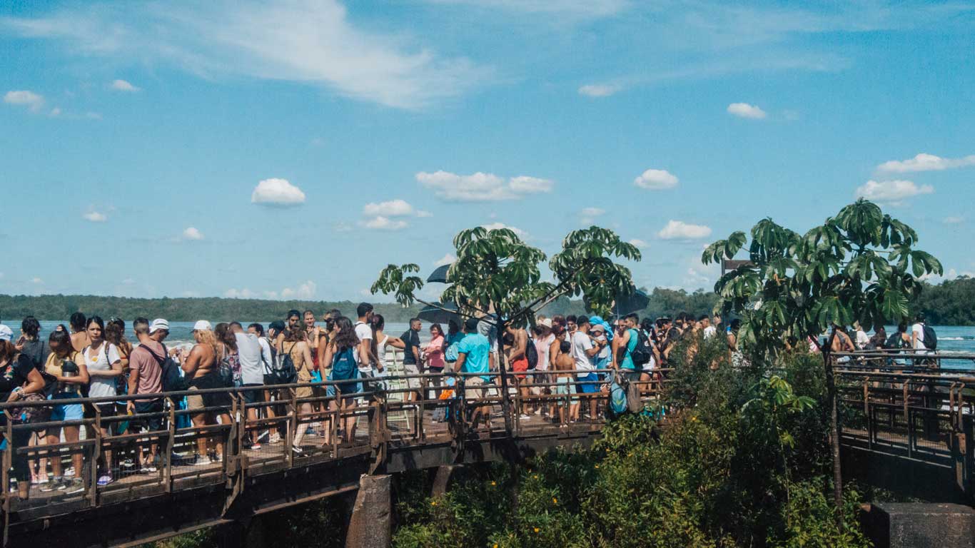 La imagen muestra una pasarela turística llena de visitantes en el parque de las Cataratas del Iguazú, con muchas personas esperando o disfrutando de las vistas bajo un cielo azul claro con algunas nubes. Al fondo, se pueden ver árboles y el río que fluye. Esta escena refleja la gran afluencia de turistas que puede encontrarse en las épocas de mayor demanda, como los meses de verano o las vacaciones escolares, lo que puede hacer menos agradable la visita debido a las multitudes.