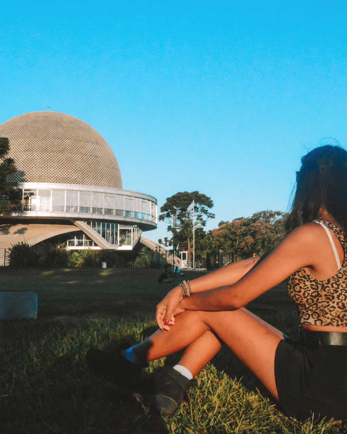 Una persona sentada en el césped frente al Planetario Galileo Galilei en Buenos Aires, Argentina. El edificio destaca por su estructura esférica, iluminada por la luz del sol, mientras el cielo azul claro complementa la escena. El entorno transmite una sensación de calma en un espacio verde y abierto.