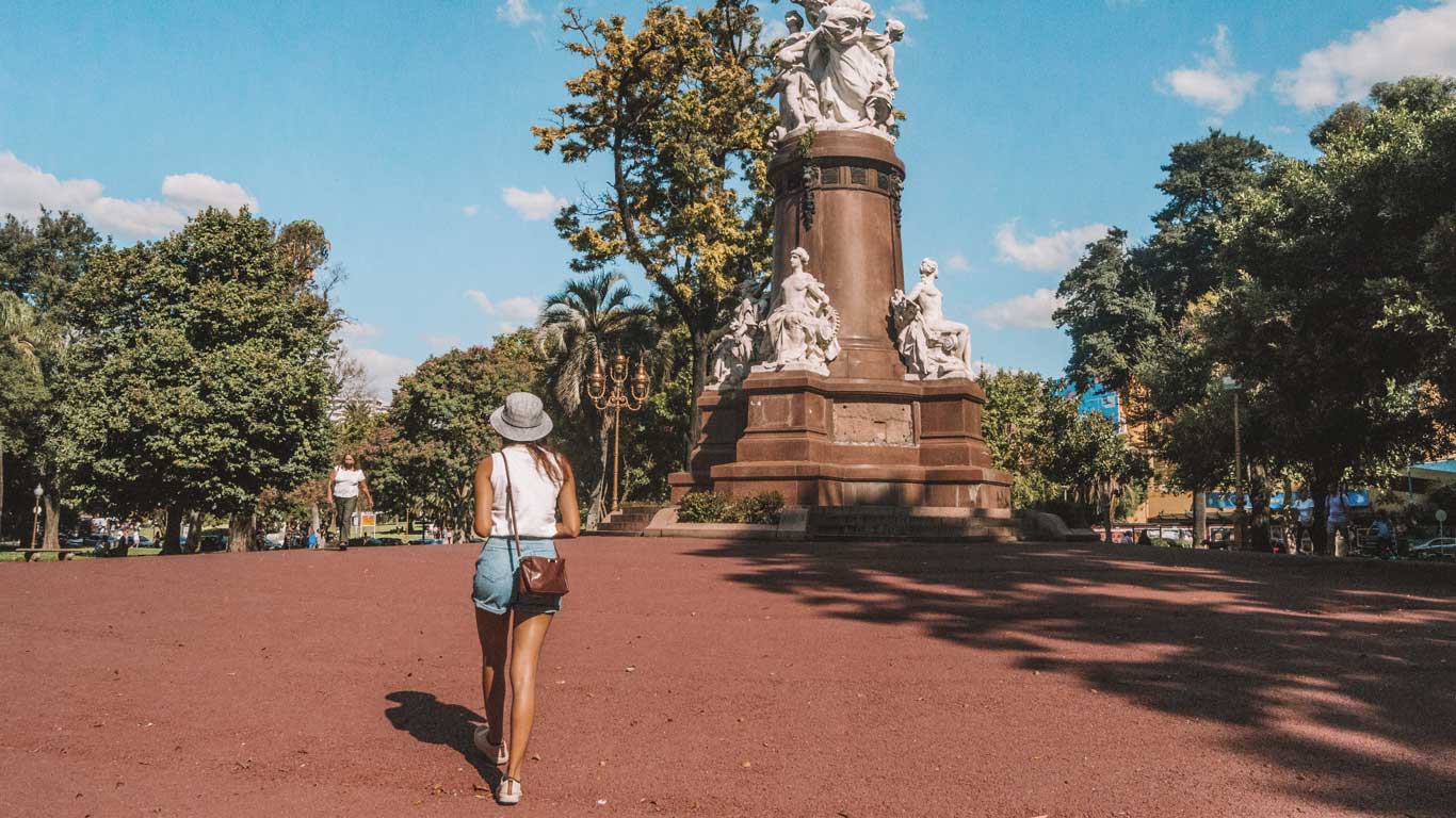 Una persona caminando hacia el monumento en la Plaza Francia, en Buenos Aires, Argentina. El monumento se destaca por sus estatuas blancas y su base elevada, rodeado de árboles y un cielo despejado. El ambiente es tranquilo, con algunas personas disfrutando del parque a la distancia.