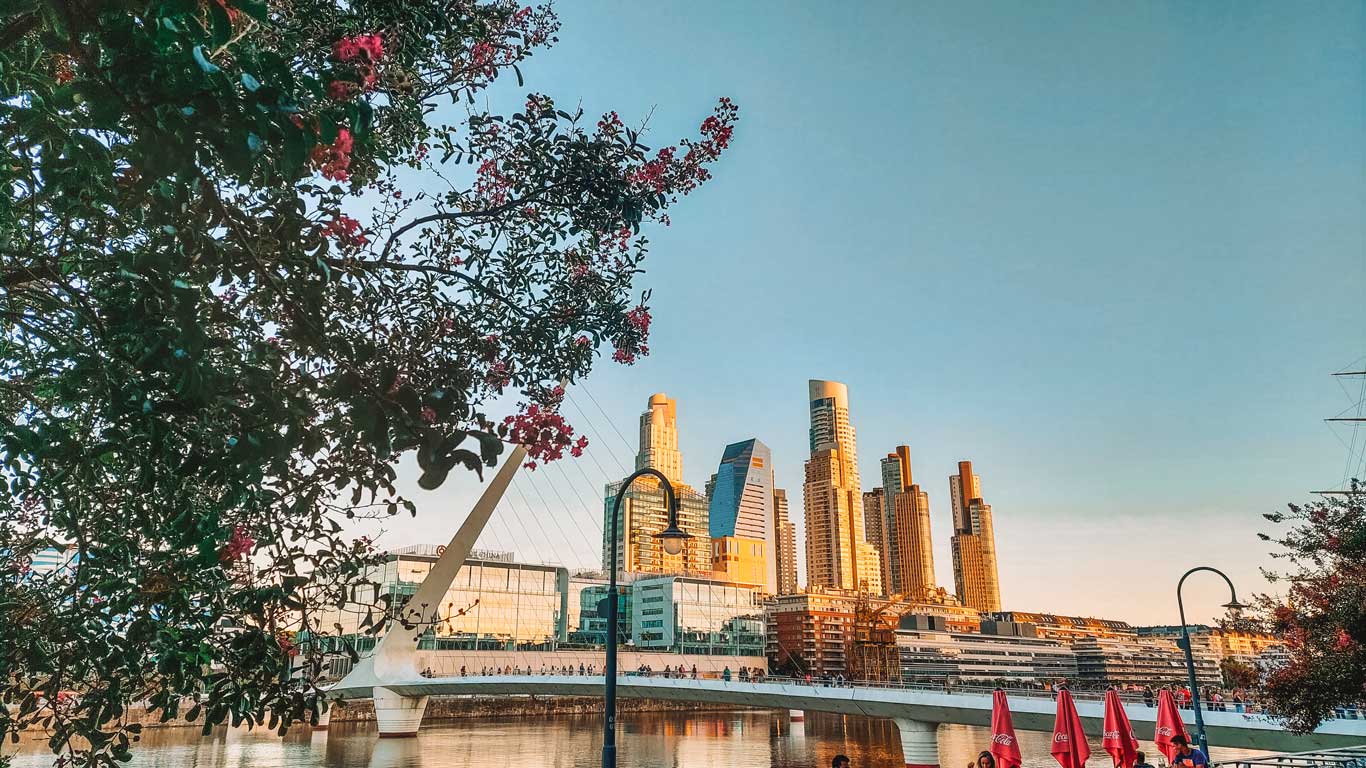 Imagen del Puente de la Mujer en Puerto Madero, Buenos Aires, Argentina, al atardecer. Se observa el reflejo del sol sobre los modernos rascacielos al fondo, mientras en primer plano hay un árbol con flores. El puente se extiende sobre el agua, rodeado por la vibrante arquitectura del distrito.