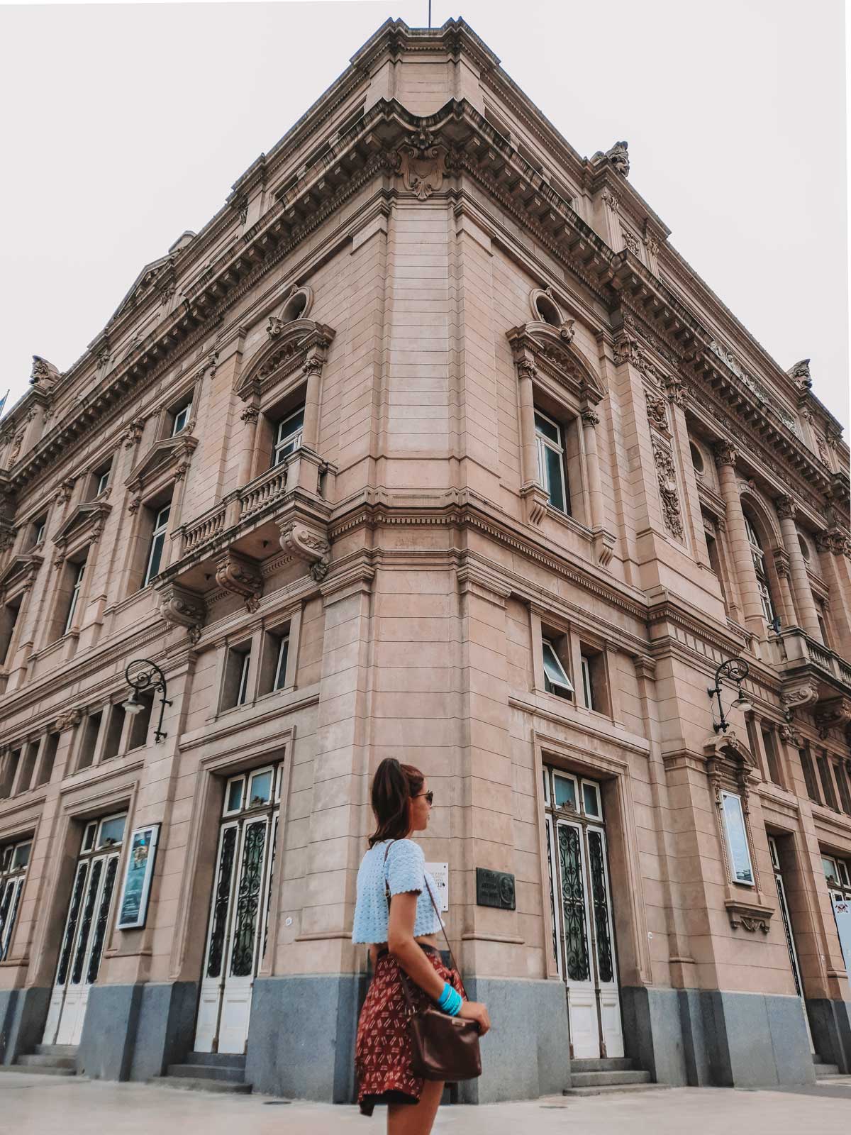 Una persona de pie, mirando hacia el majestuoso edificio del Teatro Colón en Buenos Aires, Argentina. El edificio se destaca por su arquitectura clásica y detalles ornamentales en la fachada, con ventanas y balcones decorativos que resaltan su elegancia. El cielo nublado resalta la estructura imponente del teatro.