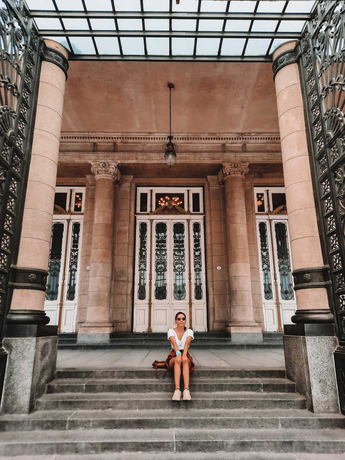 Una persona sentada en los escalones de entrada del Teatro Colón en Buenos Aires, Argentina. El fondo muestra puertas blancas con detalles decorativos y columnas clásicas bajo un techo de cristal. La escena está enmarcada por dos pilares grandes con diseños ornamentales de hierro.