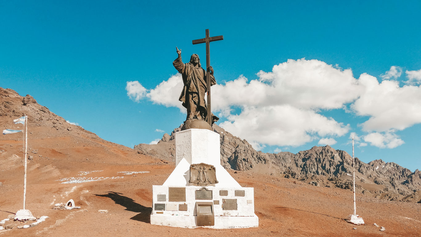 Una estatua imponente de Cristo sosteniendo una cruz, ubicada en un pedestal blanco con placas conmemorativas, rodeada por un paisaje árido de montaña en los Andes. En el fondo, picos rocosos y un cielo azul claro con algunas nubes añaden profundidad a la escena. Dos banderas argentinas ondean a los lados, destacando el carácter histórico y espiritual del lugar.