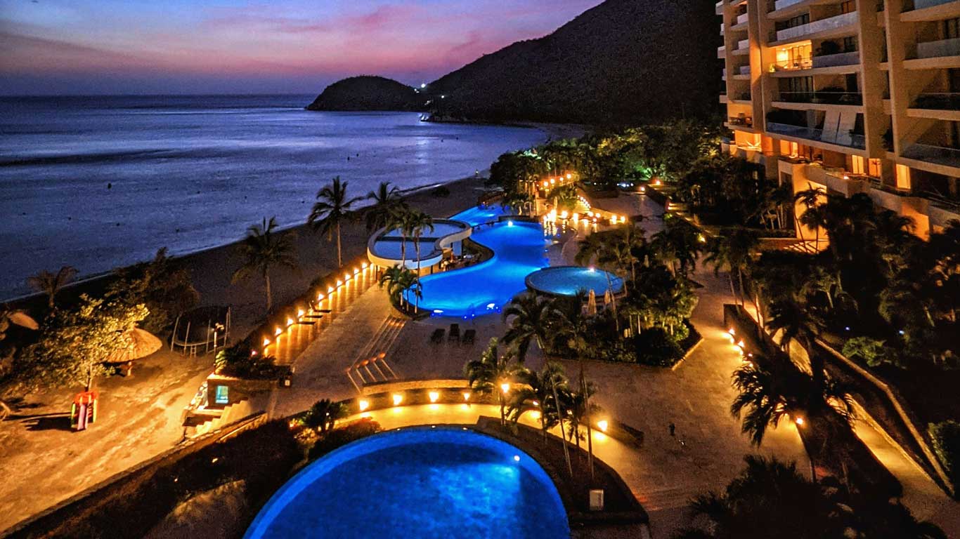 Imagen de un complejo turístico iluminado al anochecer en la costa, con piscinas curvas de agua azul brillante, rodeadas de palmeras y caminos bordeados de luces cálidas. En el fondo, se observa una playa tranquila junto al mar y colinas oscuras bajo un cielo en tonos rosados y morados. A la derecha, se ven edificios modernos con balcones iluminados.