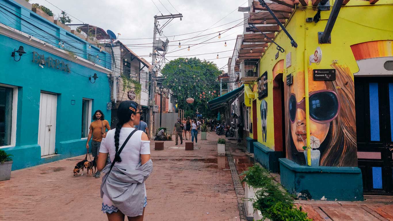 Calle peatonal en el casco histórico de Santa Marta, con edificios coloridos adornados con murales vibrantes, como el retrato de una mujer con gafas de sol. Personas pasean tranquilamente entre tiendas y cafés, mientras una enredadera de luces y un árbol frondoso decoran el ambiente. El suelo de ladrillo mojado refleja el encanto pintoresco de esta zona histórica.