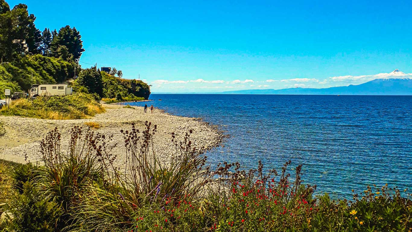 Imagen que muestra una playa de piedras en la Costanera de Puerto Varas, con vegetación en primer plano y un camino donde hay un vehículo estacionado. Al fondo, el agua azul del lago y un volcán nevado en la distancia completan el paisaje bajo un cielo despejado.