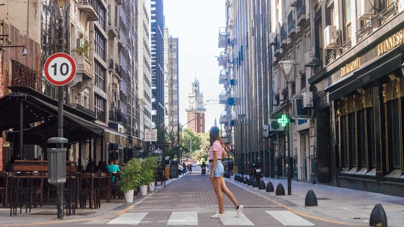 La imagen muestra una calle peatonal de Buenos Aires, Argentina, bordeada por edificios altos con balcones y fachadas antiguas. En el fondo, se puede observar la Torre de los Ingleses, un ícono de la ciudad. En primer plano, una mujer con camiseta de rayas y shorts cruza la calle, mientras a los lados hay cafés y restaurantes con mesas al aire libre.