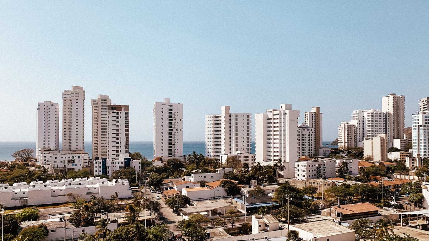 Vista panorámica del barrio El Rodadero con edificios altos y modernos frente al mar, en contraste con casas bajas rodeadas de árboles y calles tranquilas. El océano azul se extiende en el horizonte, mientras el cielo despejado resalta la arquitectura blanca de la zona. Este barrio combina un ambiente urbano y costero característico de El Rodadero, una excelente zona dónde alojarse en Santa Marta.