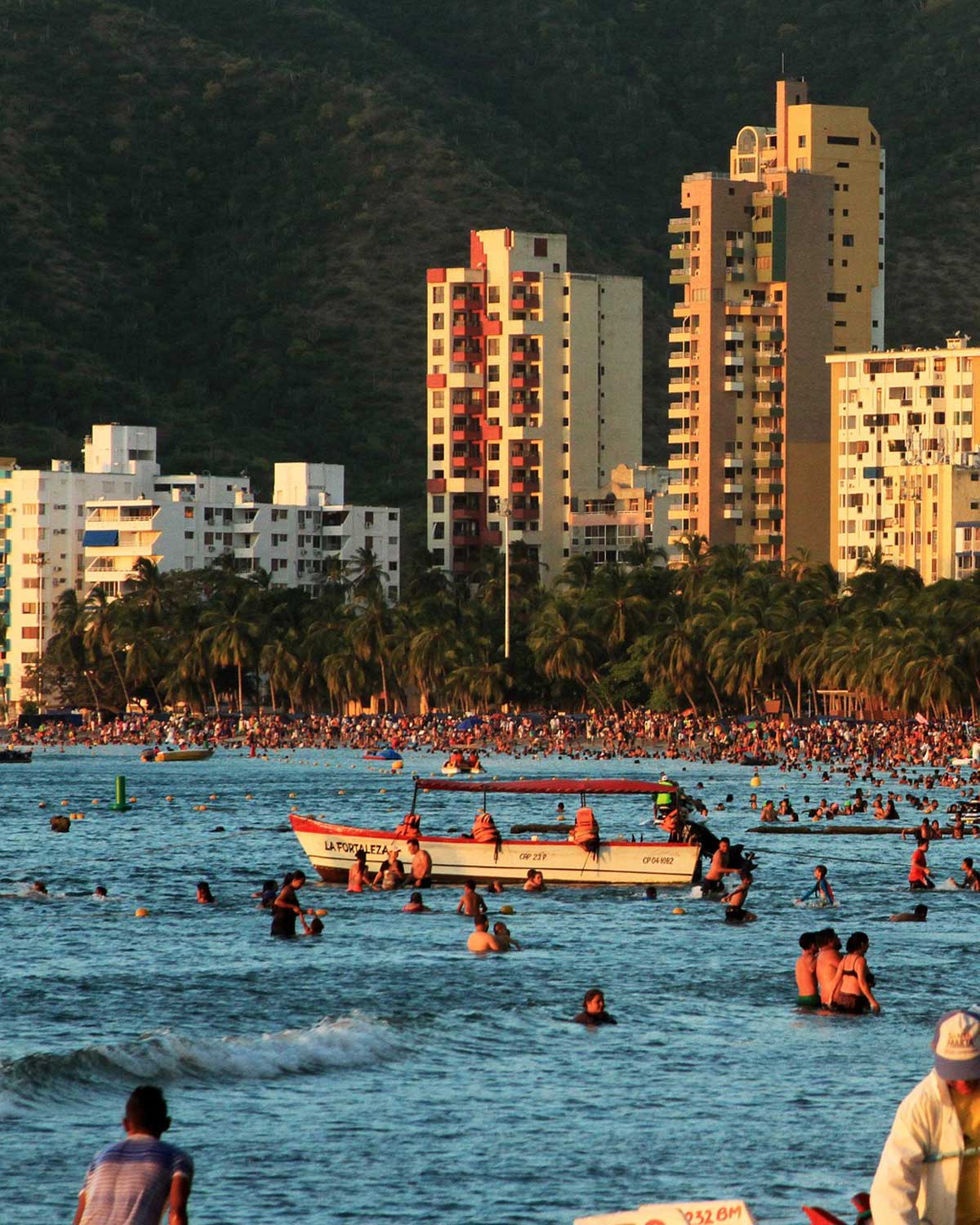 Imagen del barrio El Rodadero al atardecer, con edificios altos de apartamentos y hoteles modernos rodeados de palmeras frente a la playa. En el agua, personas nadan y disfrutan del mar junto a una lancha llamada "La Fortaleza," mientras la costa está llena de bañistas. Las montañas verdes en el fondo añaden un contraste natural a la escena urbana.