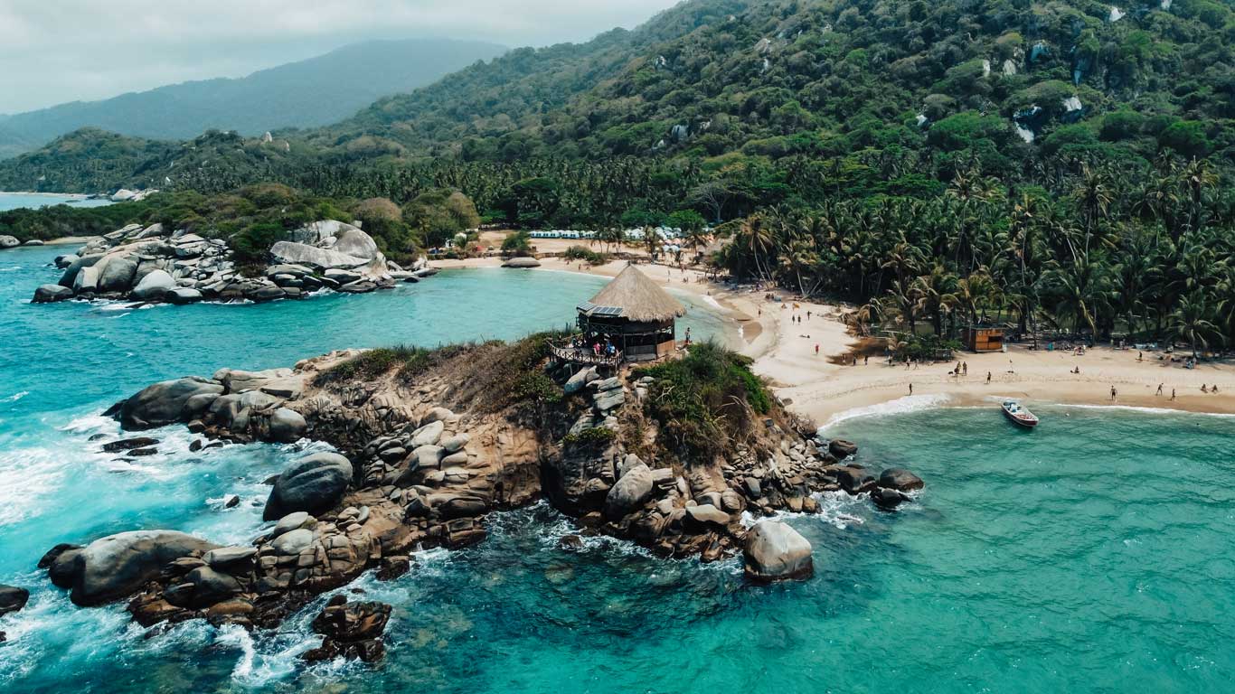 Vista aérea del Parque Nacional Tayrona, mostrando una playa paradisíaca con aguas turquesas rodeadas de palmeras y vegetación tropical. En el centro, un cabo rocoso sobresale al mar, coronado por una cabaña de techo de palma que ofrece vistas espectaculares. El paisaje combina naturaleza exuberante, arena dorada y el contraste vibrante del mar cristalino.