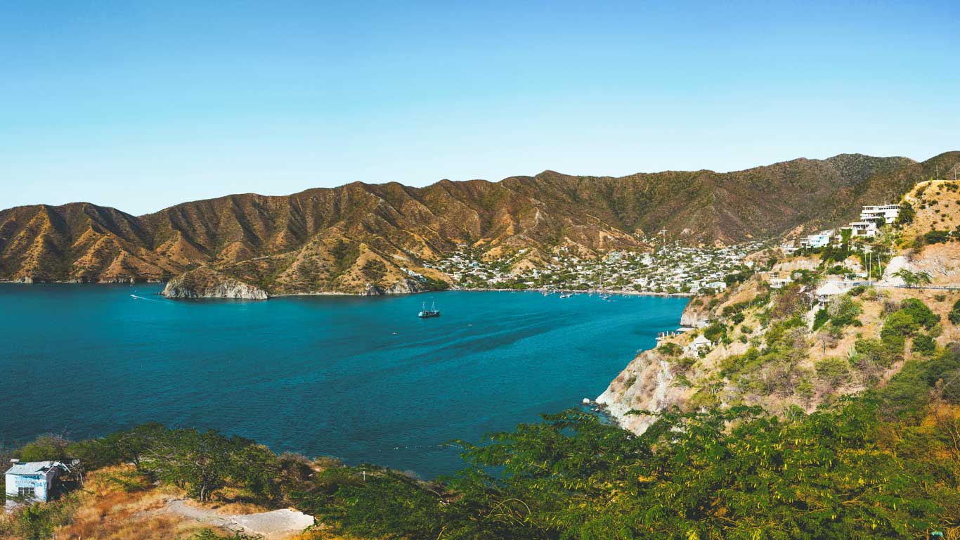 Vista panorámica de la bahía de Taganga, en Santa Marta, rodeada de montañas áridas con tonos dorados y verdes. El mar azul profundo alberga pequeños barcos y veleros, mientras que en la costa se observan casas blancas dispersas en las colinas. El paisaje combina belleza natural y tranquilidad costera bajo un cielo despejado.