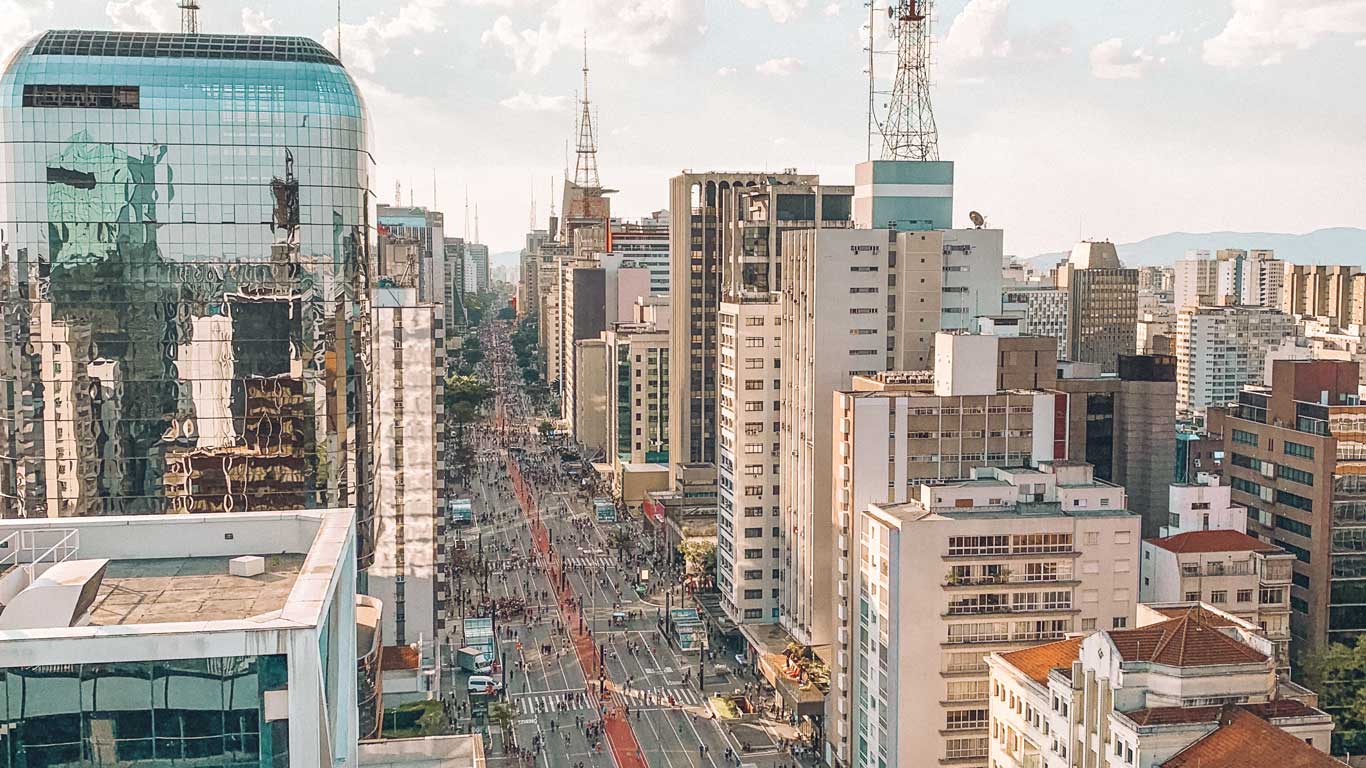 Esta imagen muestra una vista aérea de una avenida Paulista, São Paulo, rodeada de altos edificios modernos, algunos con fachadas de vidrio que reflejan el cielo. En el centro, la avenida, posiblemente con carriles exclusivos para autobuses, está llena de personas y actividad, sugiriendo un ambiente vibrante en una gran ciudad. Al fondo, se observan más edificios y antenas que destacan en el horizonte.