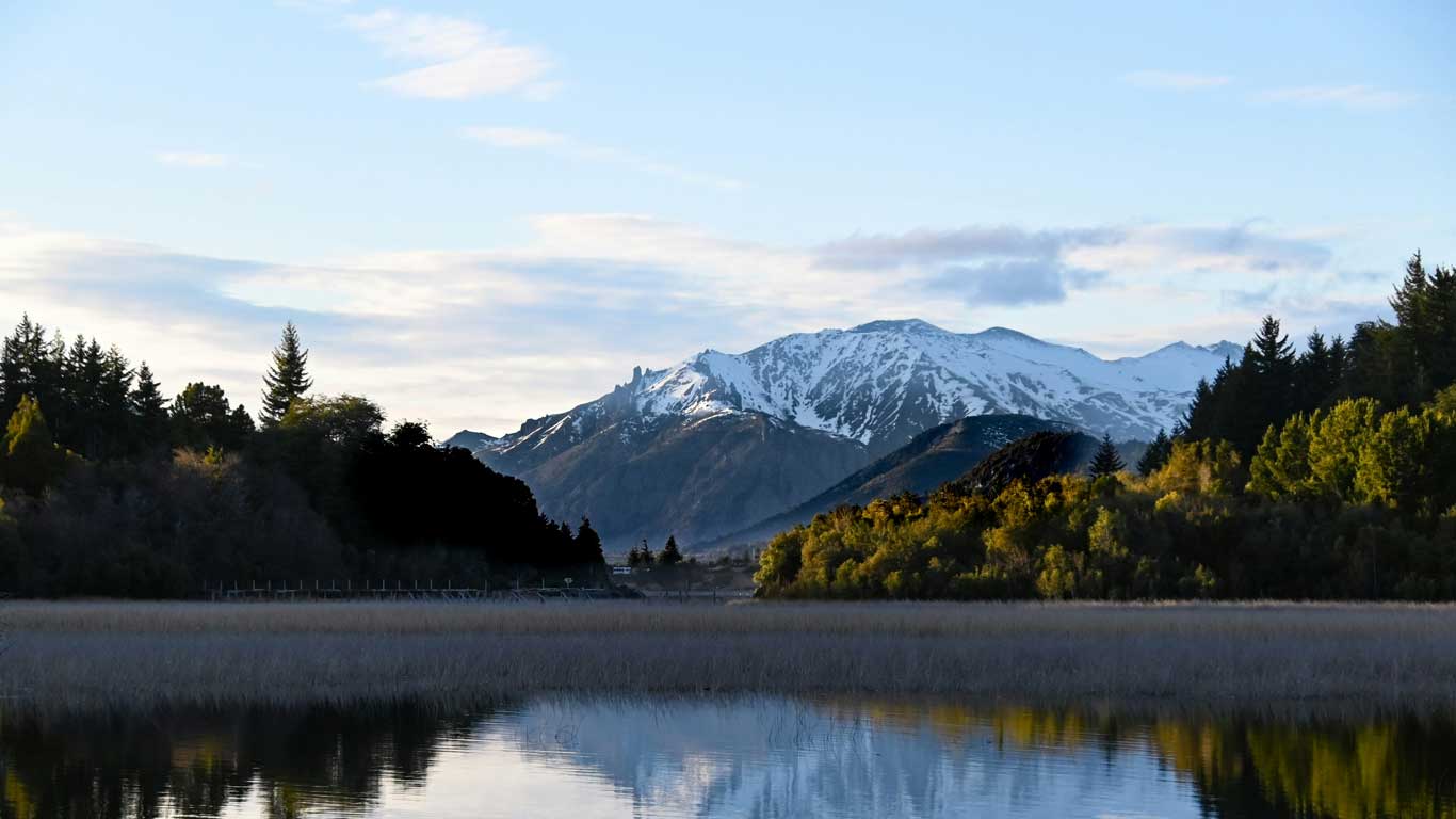 Imagen que muestra un paisaje en Bariloche, Argentina, con un lago tranquilo rodeado de vegetación frondosa. Al fondo, se destacan montañas cubiertas de nieve bajo un cielo parcialmente nublado, creando una escena serena y pintoresca. Este lugar es ideal para actividades al aire libre como senderismo, fotografía y disfrutar de la naturaleza en su máxima expresión.