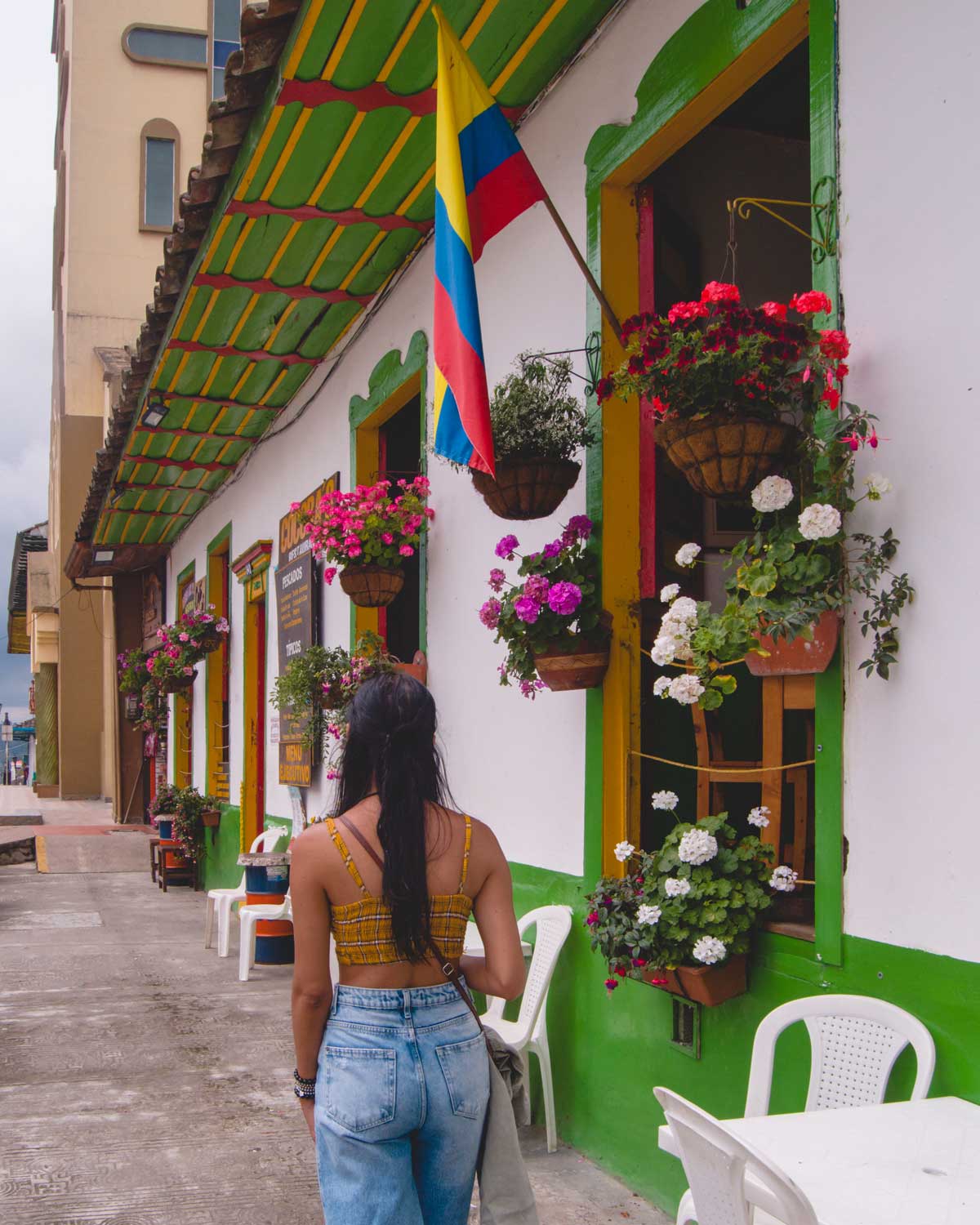 Una mujer con un top amarillo y pantalones vaqueros camina por una acera frente a una casa típica de Salento, Colombia. Las paredes blancas con detalles verdes y ventanas adornadas con coloridas flores en macetas colgantes, junto con una bandera colombiana ondeando, destacan en la fachada. El ambiente es acogedor y pintoresco, representando el encanto del centro de este pueblo cafetero.