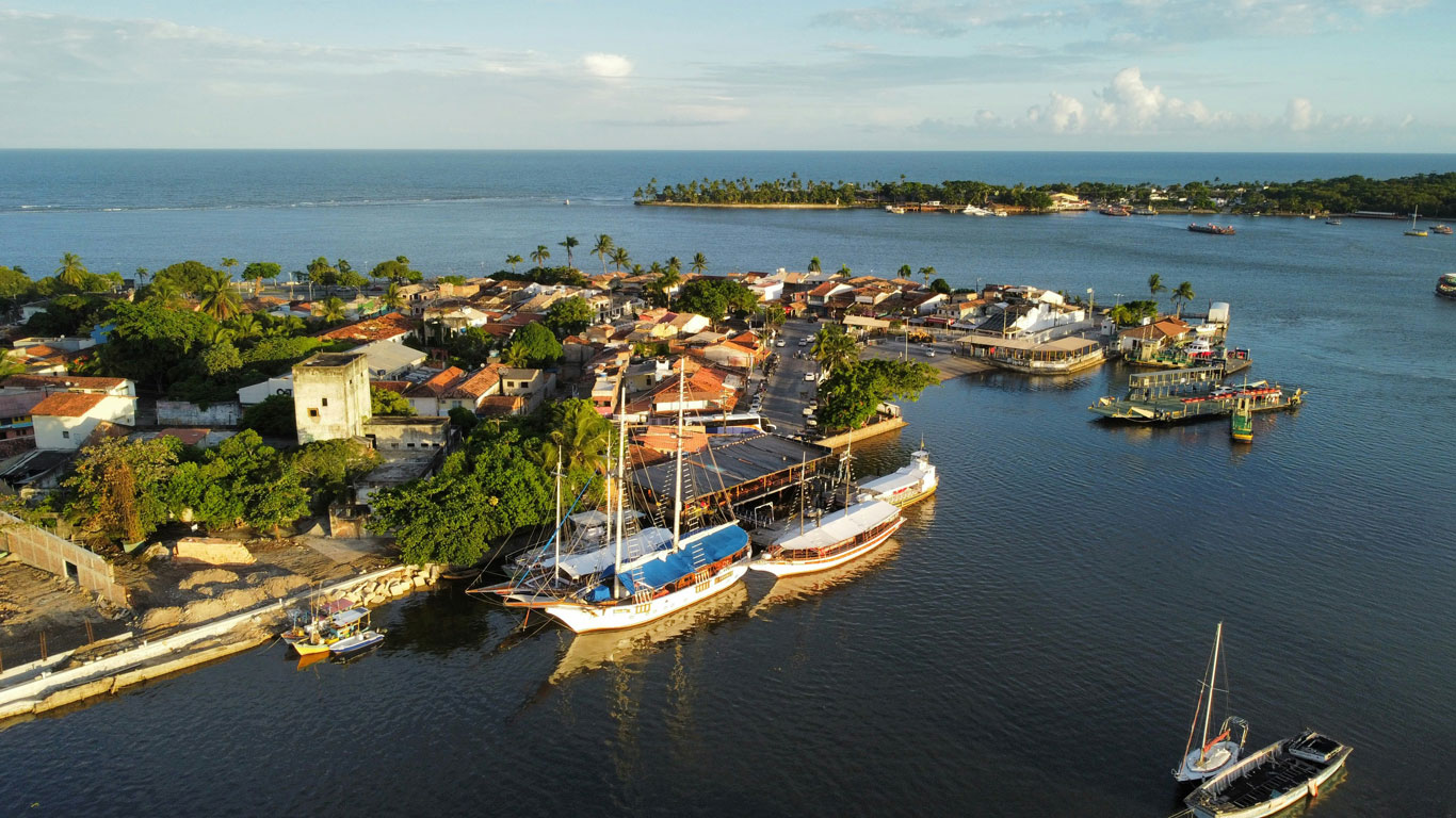 Vista aérea del centro histórico de Porto Seguro, Brasil, que muestra casas coloridas con techos de tejas rojas rodeadas de una exuberante vegetación. Varias embarcaciones, incluido un velero con cubiertas azules, están atracadas a lo largo de la costa, mientras que las tranquilas aguas reflejan el cálido resplandor del sol poniente. Al fondo, una franja de tierra bordeada de palmeras se extiende hacia el océano bajo un cielo despejado.