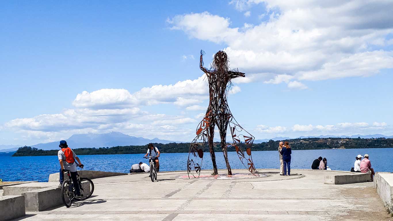 Imagen que muestra una escultura de metal en forma humana situada en la Costanera de Puerto Varas, frente a un lago azul bajo un cielo despejado con algunas nubes. A su alrededor, se observan personas caminando, sentadas y disfrutando del paisaje, incluyendo ciclistas. Al fondo, se aprecian montañas y vegetación, creando un ambiente relajante y natural.