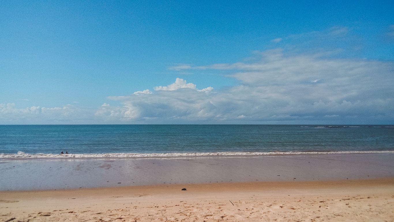 Playa de Curuípe en Porto Seguro, Brasil, con una tranquila franja de arena dorada y el mar azul extendiéndose hasta el horizonte. Las suaves olas tocan la orilla mientras dos personas disfrutan del agua en la distancia. El cielo presenta una mezcla de azul claro con nubes dispersas, creando una atmósfera relajante y serena.