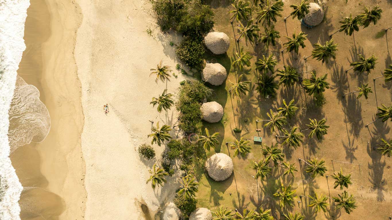 Vista aérea de una playa en el Parque Tayrona, con arenas doradas, olas suaves y cabañas tradicionales rodeadas de palmeras. La imagen muestra una combinación de naturaleza virgen y alojamiento rústico, ideal para quienes buscan dónde alojarse en el Parque Tayrona.