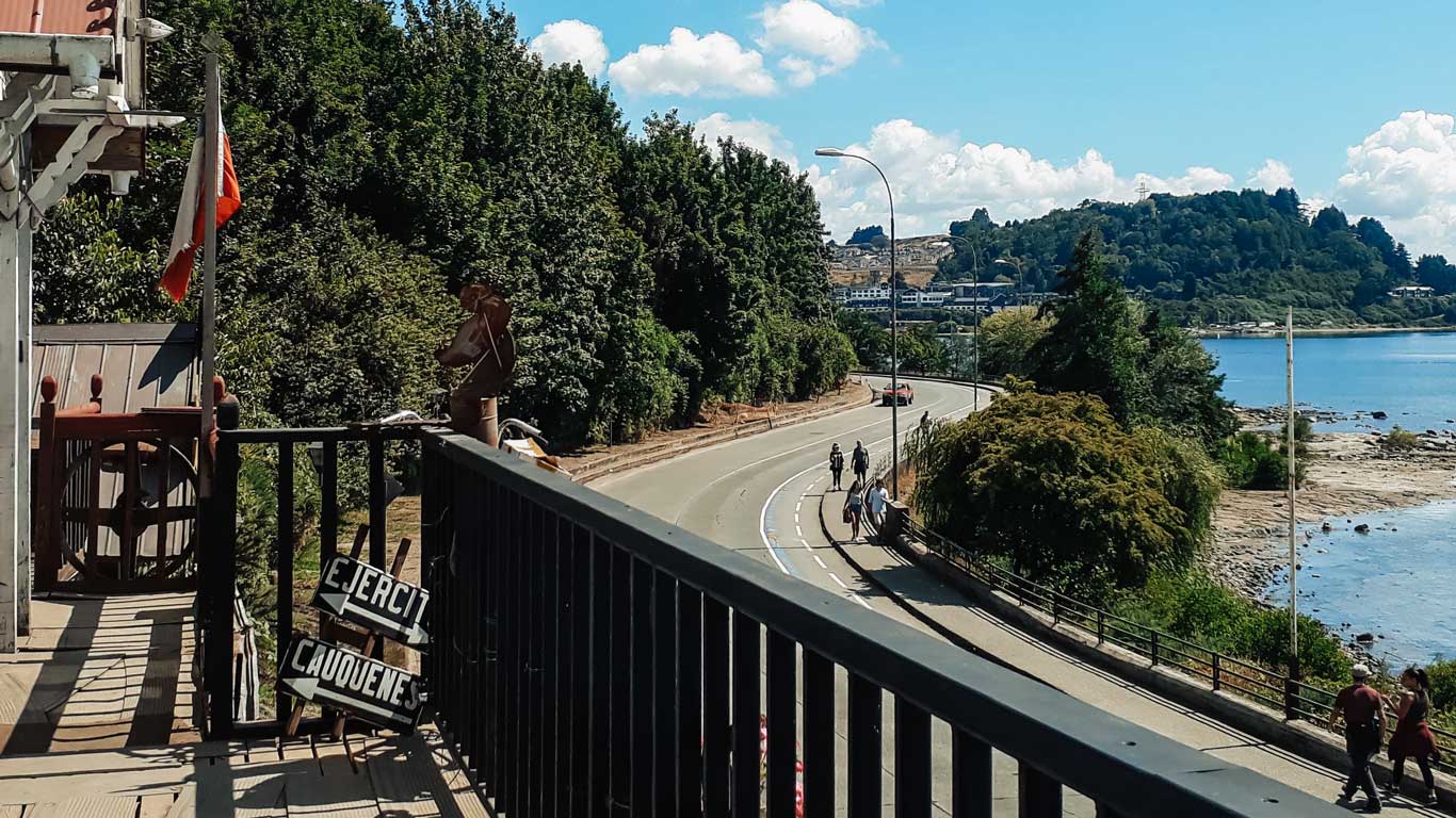 Una vista desde un balcón en Puerto Varas, Chile, con una carretera curva bordeada por árboles frondosos y un sendero junto al lago. Una excelente zona donde alojarse en Puerto Varas. Al fondo, se observan colinas y edificaciones, mientras el cielo despejado y el agua cristalina completan el paisaje. La bandera chilena ondea en el balcón, resaltando el ambiente tranquilo y pintoresco del lugar.