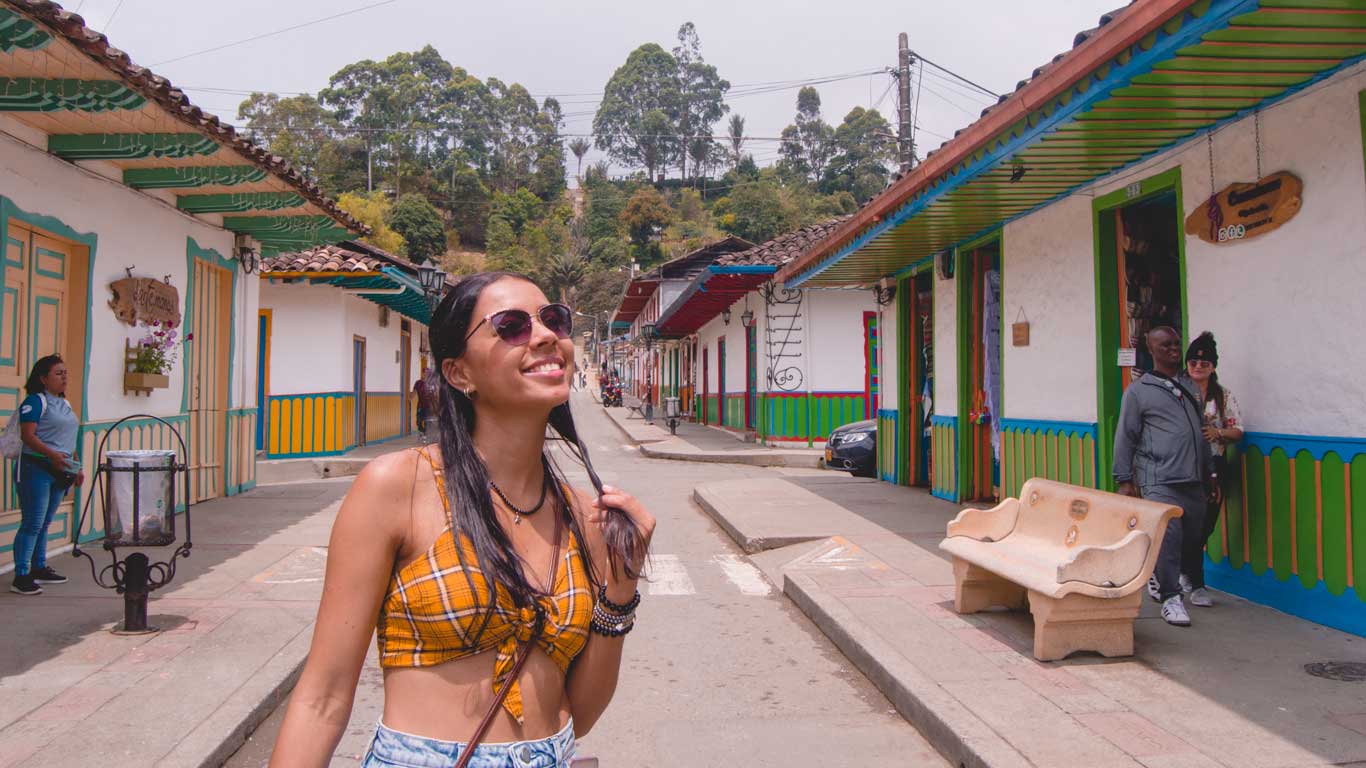 Una mujer sonriente con gafas de sol y un top amarillo a cuadros camina por una colorida calle de Salento, Colombia. Las casas están pintadas de blanco con detalles verdes, amarillos y azules, típicos de la arquitectura de la región cafetera. El ambiente es alegre, con personas disfrutando del día y un paisaje verde al fondo que enmarca el lugar.