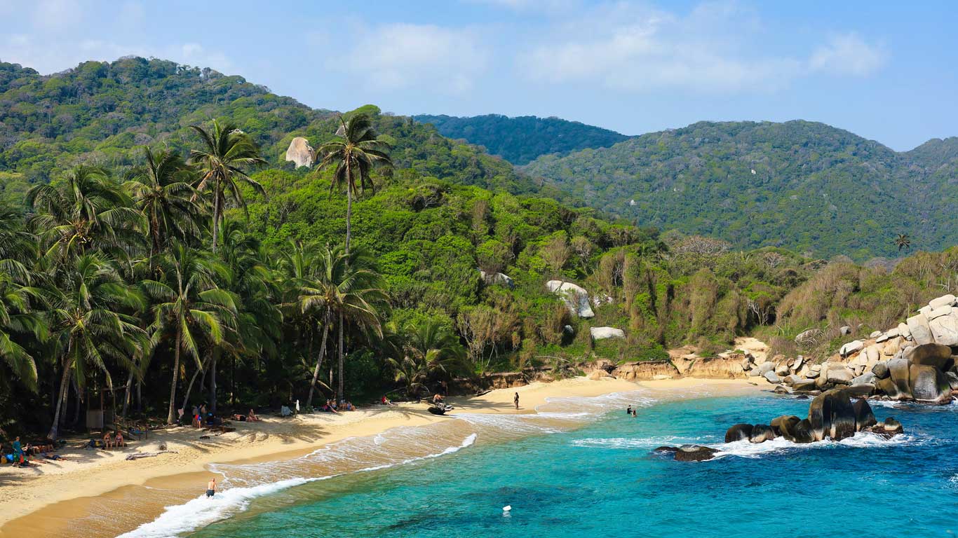 Playa rodeada de exuberante vegetación y aguas cristalinas en el Parque Tayrona, con turistas disfrutando del sol y el paisaje natural. 