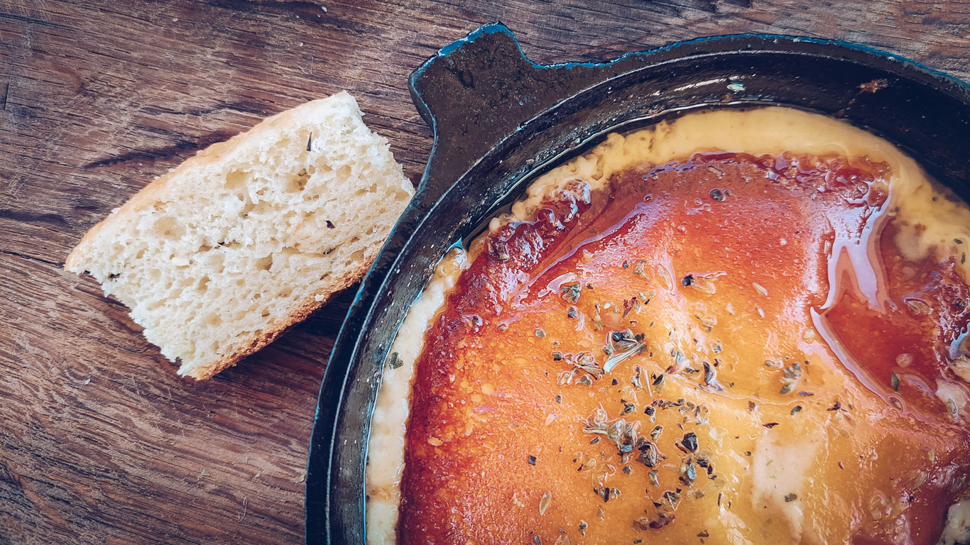 Una sartén de hierro con una provoleta derretida, dorada y cubierta de orégano, junto a un trozo de pan artesanal sobre una mesa de madera. La provoleta tiene una textura crujiente en la parte superior y el queso fundido se desborda ligeramente por los bordes. Una comida típica argentina, perfecta para acompañar con pan y disfrutar en una mesa rústica.