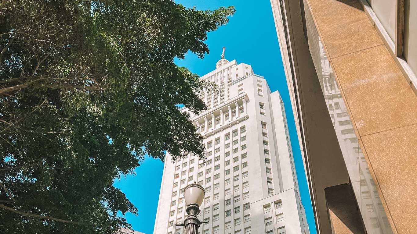 La imagen muestra un edificio alto y blanco con arquitectura moderna y ventanas alineadas, conocido como el Farol Santander, situado en São Paulo. El cielo azul y los árboles en primer plano enmarcan la estructura, que se destaca por su diseño imponente y simbólico. La escena combina la arquitectura urbana con elementos naturales, creando un contraste visual atractivo.