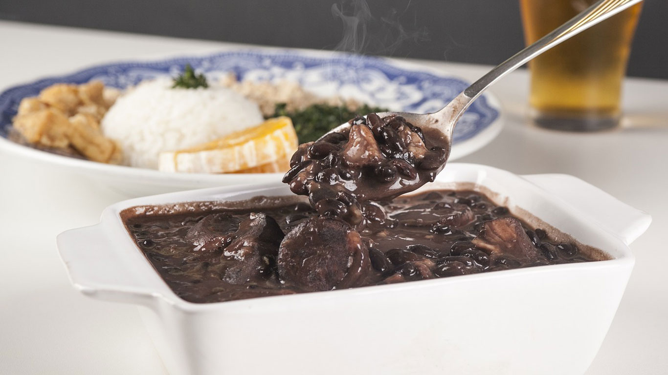 La imagen muestra un plato típico brasileño llamado feijoada, servido en una fuente blanca y acompañado de guarniciones como arroz blanco, naranjas en rodajas, col rizada y farofa al fondo. La feijoada está humeante, con su característico caldo espeso y trozos de carne y embutidos, resaltando su aspecto apetitoso. Este platillo es una especialidad tradicional de la cocina brasileña, ideal para disfrutar en un almuerzo abundante.