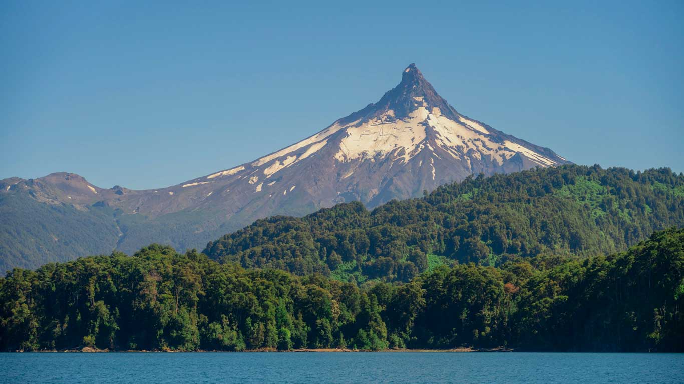 Imagen que muestra el majestuoso volcán Puntiagudo en Chile, con su característica cima afilada parcialmente cubierta de nieve, rodeado por una exuberante vegetación verde. En el primer plano, el lago de Todos los Santos resalta con su agua cristalina bajo un cielo despejado. 