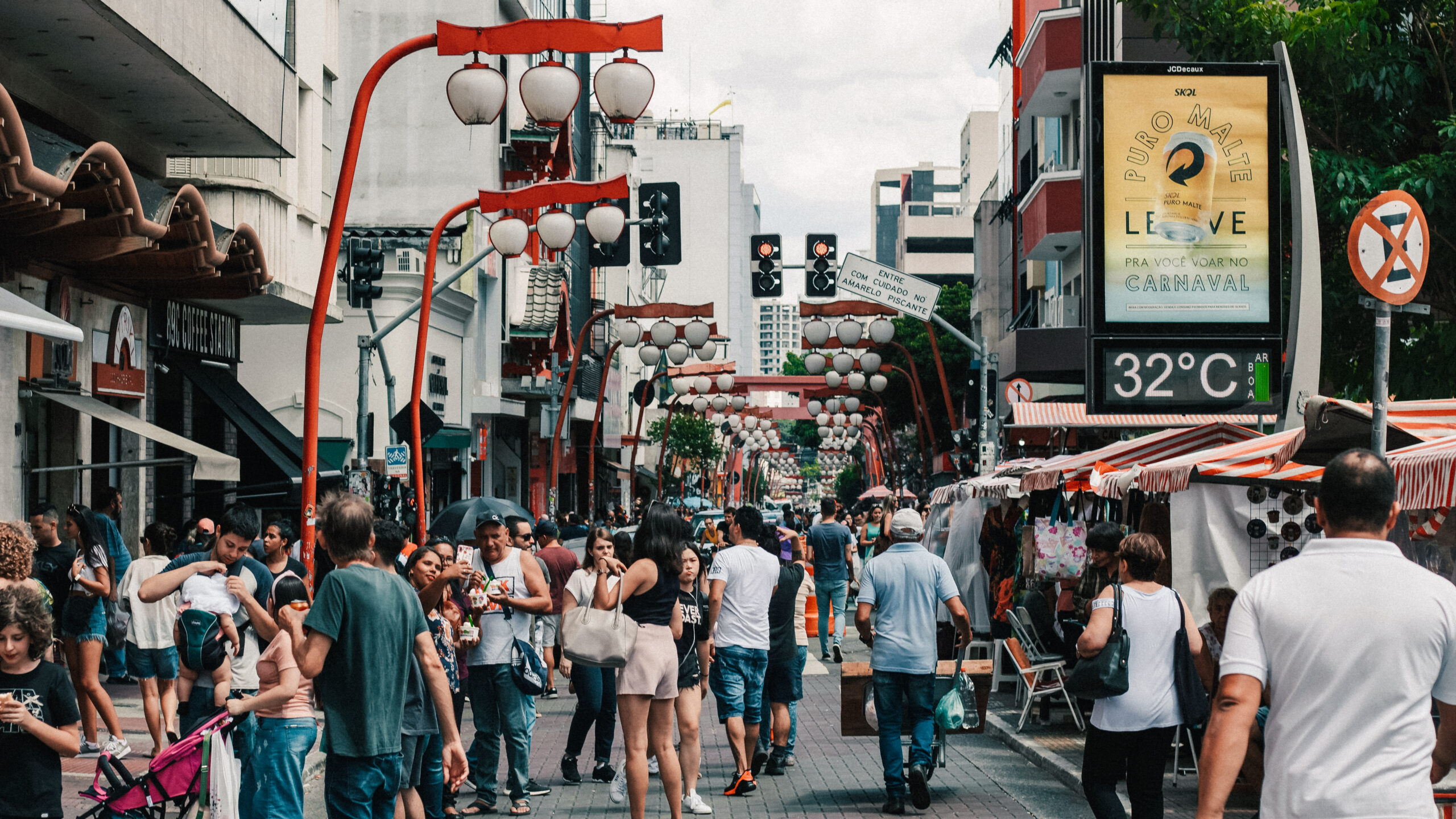 La imagen muestra una concurrida calle del barrio Liberdade en São Paulo, decorada con faroles tradicionales japoneses y estructuras rojas características de la cultura asiática. La zona está llena de personas caminando entre los puestos del mercado, mientras tiendas y edificios modernos se alinean a ambos lados. Este lugar emblemático combina la vida urbana con la herencia cultural japonesa, creando un ambiente vibrante y multicultural.