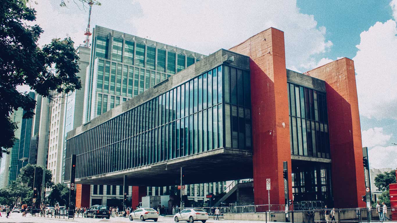 La imagen muestra el edificio del MASP (Museo de Arte de São Paulo), una estructura icónica con un diseño moderno y minimalista. El edificio está suspendido sobre pilares rojos que lo elevan, con amplios ventanales de vidrio que reflejan el cielo y los alrededores urbanos. Este lugar es un importante centro cultural y artístico en São Paulo, conocido por su arquitectura única y sus exposiciones.