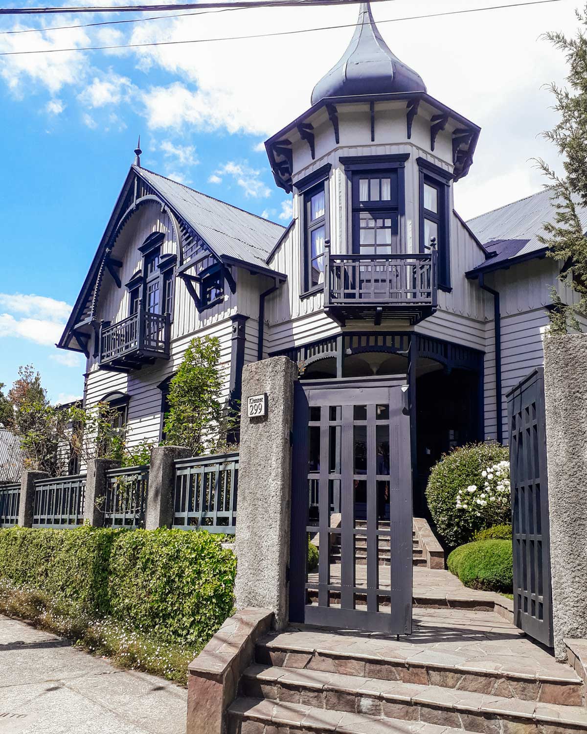 Una casa de estilo arquitectónico tradicional alemán en Puerto Varas, Chile, con paredes blancas, detalles en negro y un tejado inclinado con una torre distintiva. El jardín bien cuidado rodea la propiedad, con arbustos verdes y flores blancas. El cielo despejado y la fachada elegante resaltan la belleza de esta construcción típica de la región.