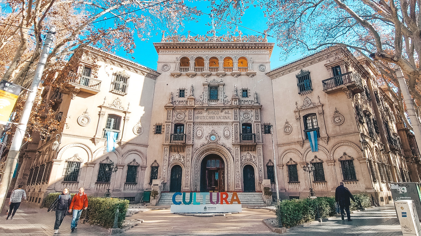 Esta imagen muestra la fachada del edificio del Banco Hipotecario Nacional en Mendoza, Argentina, con una arquitectura clásica y detallada. En el frente se encuentra un letrero colorido que dice "CULTURA", junto con una bandera argentina colgada en el centro. El cielo despejado y las hojas de los árboles en tonos otoñales completan la escena.