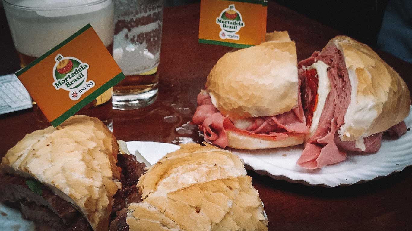 La imagen muestra dos grandes sándwiches servidos en un plato, elaborados con panes crujientes y rellenos generosos de mortadela, queso y otros ingredientes. Junto a ellos, hay un vaso de bebida y una pequeña bandera con el logo de "Mortadela Brasil", que identifica el lugar como parte de la oferta gastronómica típica del Mercado Municipal de São Paulo. La escena resalta una experiencia culinaria clásica en la ciudad.