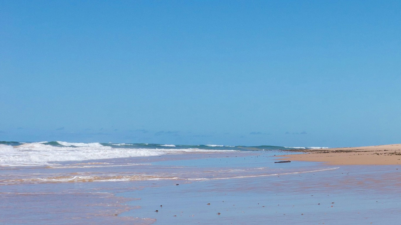 Playa de Mundaí, una de las mejores áreas dónde alojarse en Porto Seguro, Brasil, con arena dorada y suaves olas rompiendo en la orilla bajo un cielo azul despejado. La playa tranquila muestra poca presencia de personas o construcciones, ofreciendo una atmósfera serena. 